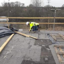 BEST HOME MARIUSZ MIKOŁAJCZYK - Pracownik w kasku i kamizelce odblaskowej, klęczący na płaskim dachu pokrytym papą, wykonuje uszczelnienia. W tle widoczne drzewa, słup energetyczny i fragment barierek.