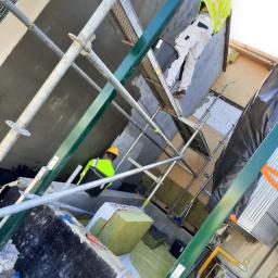 BEST HOME MARIUSZ MIKOŁAJCZYK - Pracownicy na rusztowaniu podczas nakładania warstwy szpachli na elewację budynku, widoczne materiały izolacyjne i narzędzia.