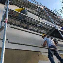 BEST HOME MARIUSZ MIKOŁAJCZYK - Pracownik w żółtym kasku na rusztowaniu, wyrównuje szpachlą fragment elewacji domu, widoczne ślady wcześniejszych warstw i fragment ocieplenia.