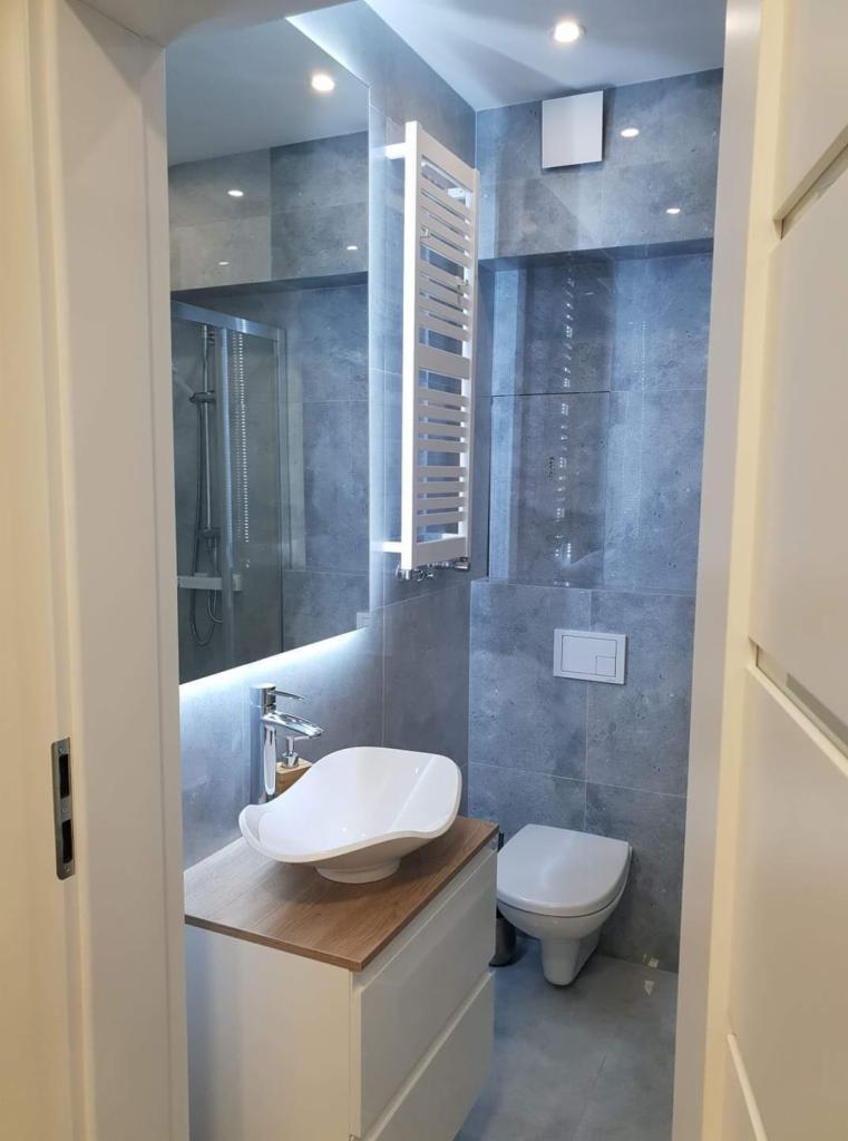 Nowoczesna łazienka z szarą glazurą, umywalką nablatową o nietypowym kształcie, podświetlanym lustrem i grzejnikiem drabinkowym.