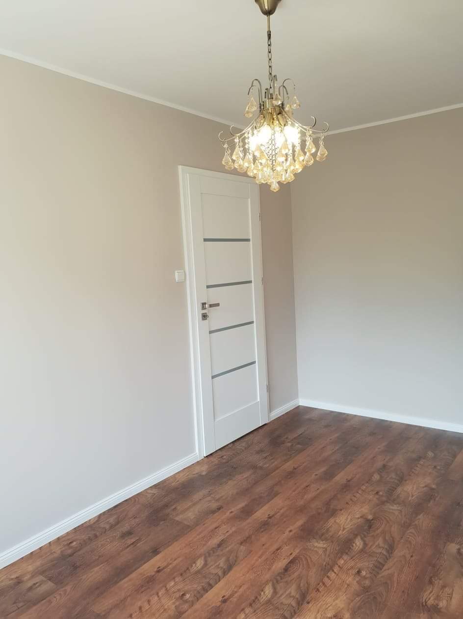 Pomalowane na beżowo wnętrze pokoju z drewnopodobną podłogą, białą, nowoczesną lampą z kryształkami i białą, minimalistyczną stolarką drzwiową z szarymi wstawkami.