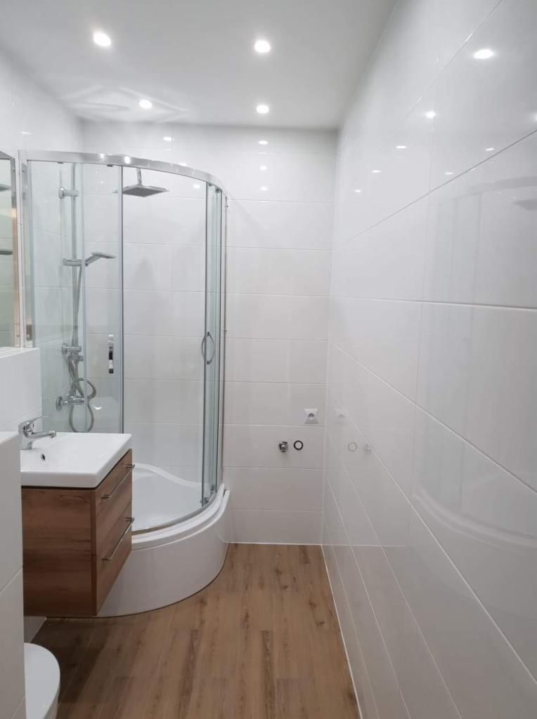 Nowoczesna, jasna łazienka po remoncie z kabiną prysznicową z zaokrąglonym frontem, umywalką z szafką z drewnopodobnym fornirem i błyszczącymi, białymi płytkami na ścianach oraz panelami...