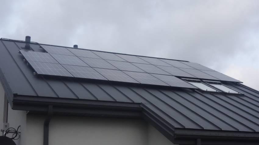 Panele słoneczne na dachu z blachy trapezowej w kolorze grafitowym, zamontowane obok okien dachowych, pochmurne niebo w tle.