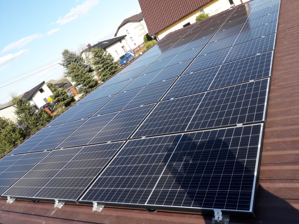 Panel słoneczny na brązowym dachu domu jednorodzinnego w słoneczny dzień, widoczne domy w tle.