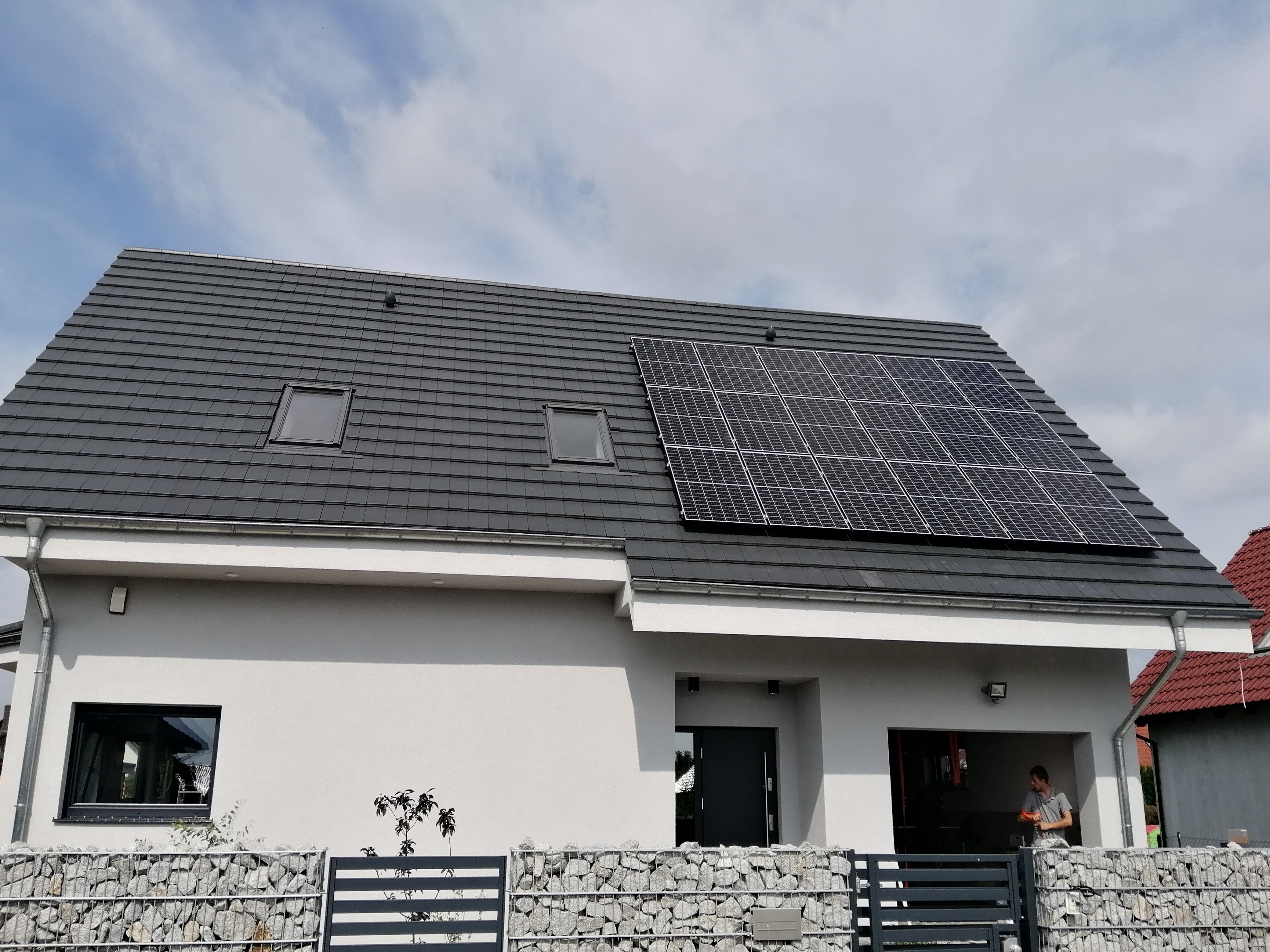 Nowoczesny dom jednorodzinny z szarą dachówką i panelami słonecznymi na dachu, widoczne dwa okna dachowe, elewacja w kolorze złamanej bieli, ogrodzenie gabionowe, w tle częściowo widoczny sąsiedni...