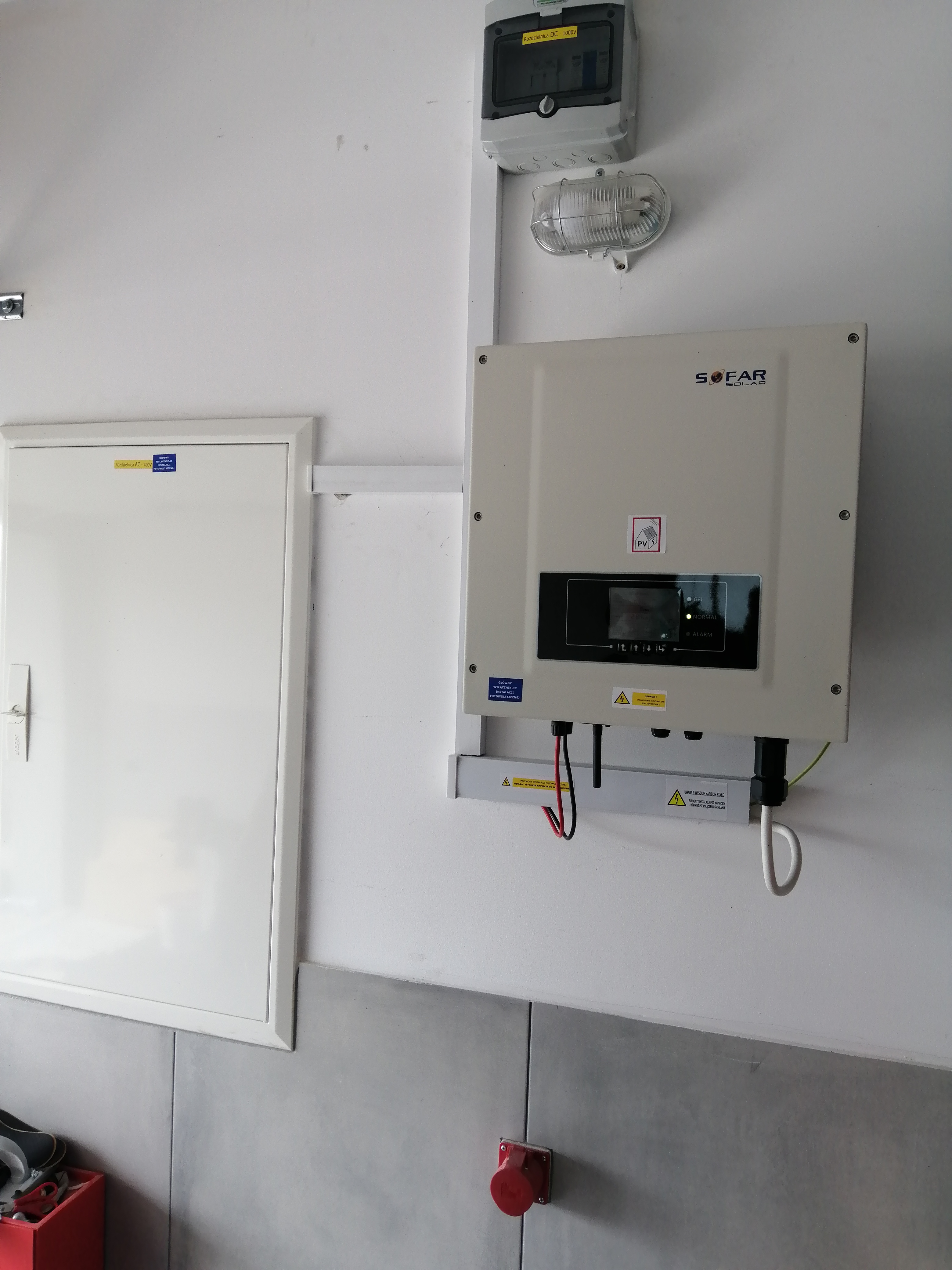 Zamontowany na ścianie inwerter Sofar Solar z widocznymi przewodami, obok skrzynka elektryczna i rozdzielnica. Tabliczki ostrzegawcze o napięciu stałym.
