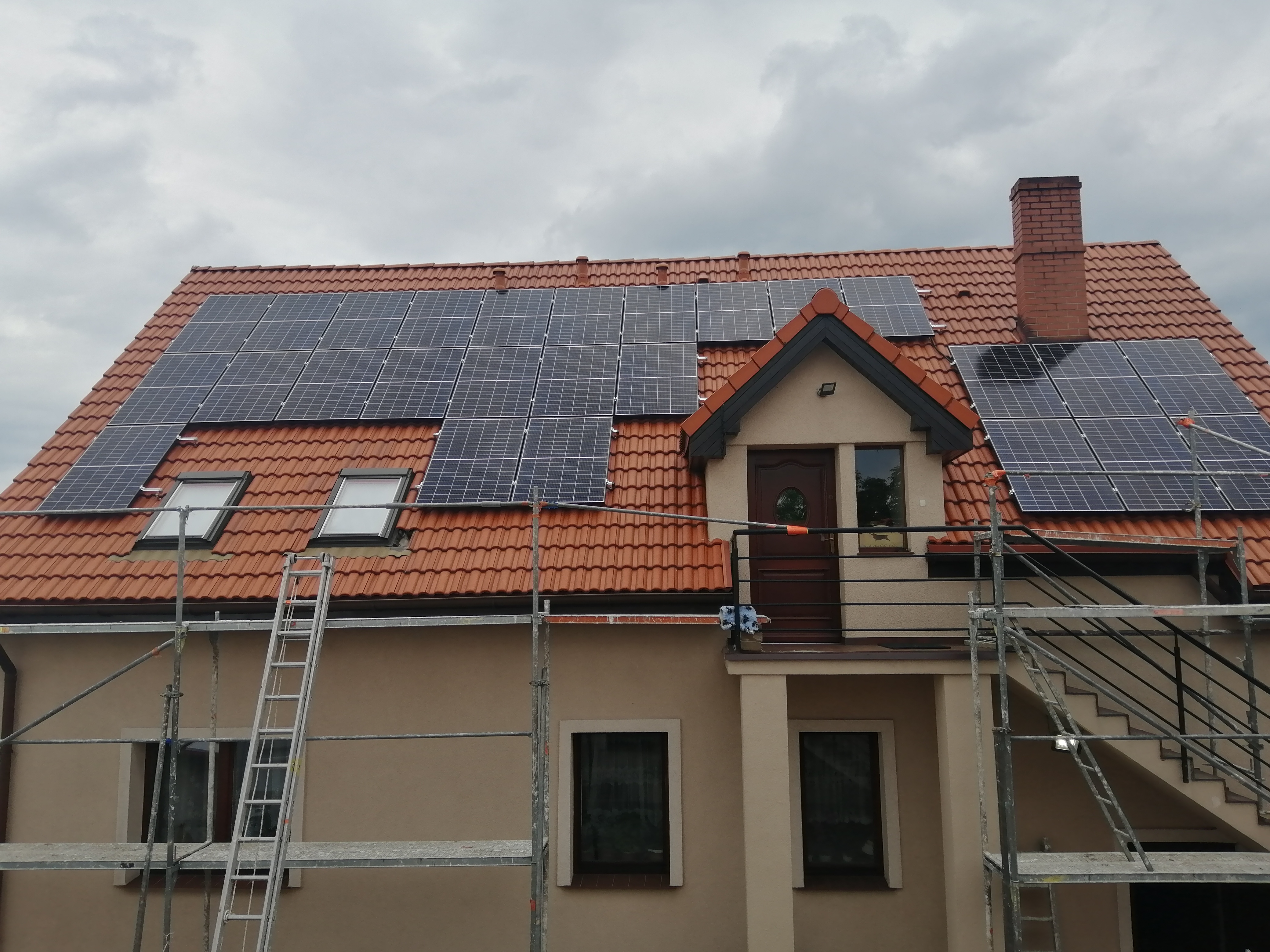 Dom z czerwoną dachówką, na której zamontowane są panele słoneczne. Widoczne rusztowania i drabina, sugerujące trwający montaż.