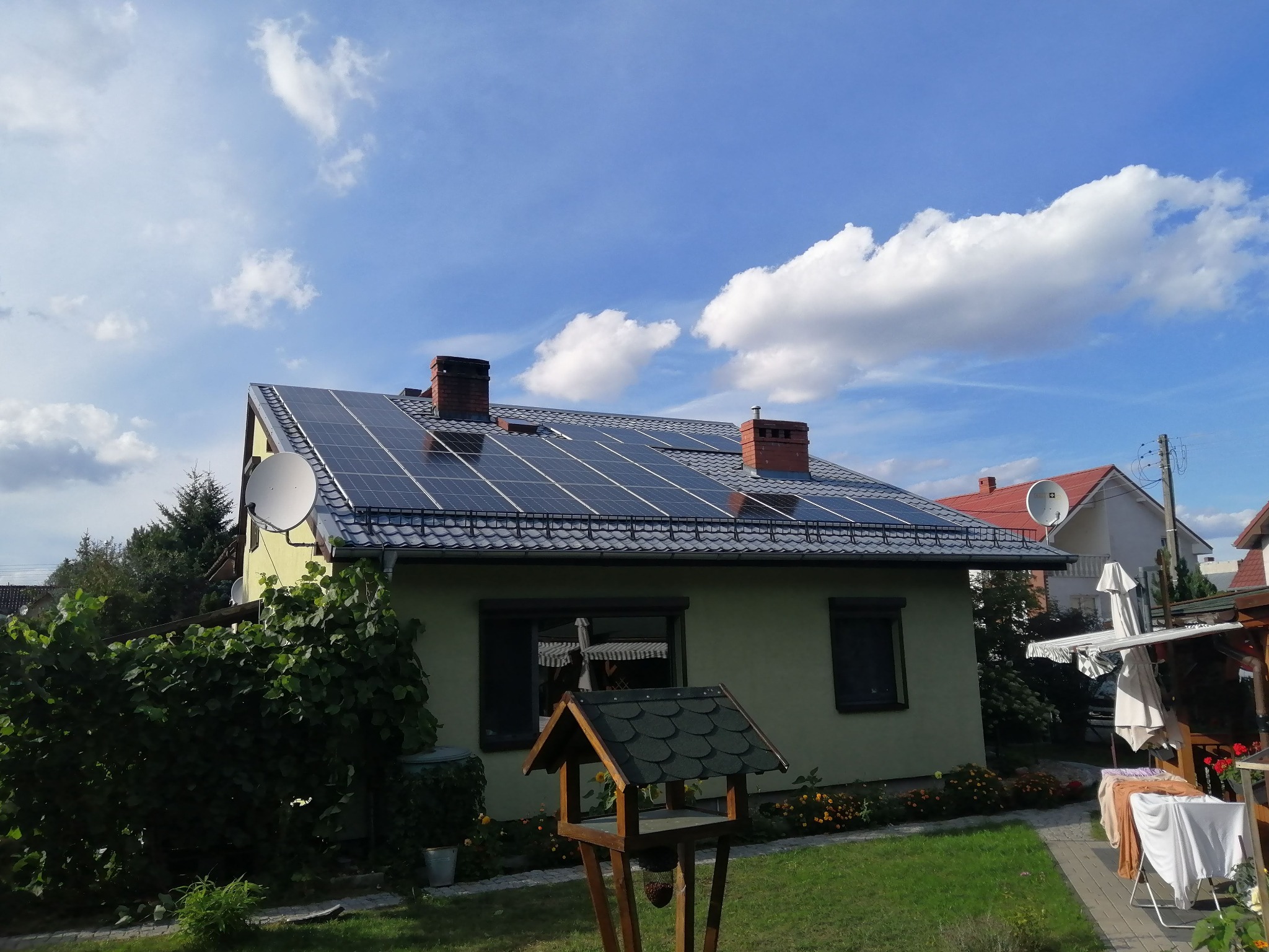 Dom jednorodzinny z panelami słonecznymi na dachu, widoczne dwa kominy i antena satelitarna, na pierwszym planie karmnik dla ptaków.
