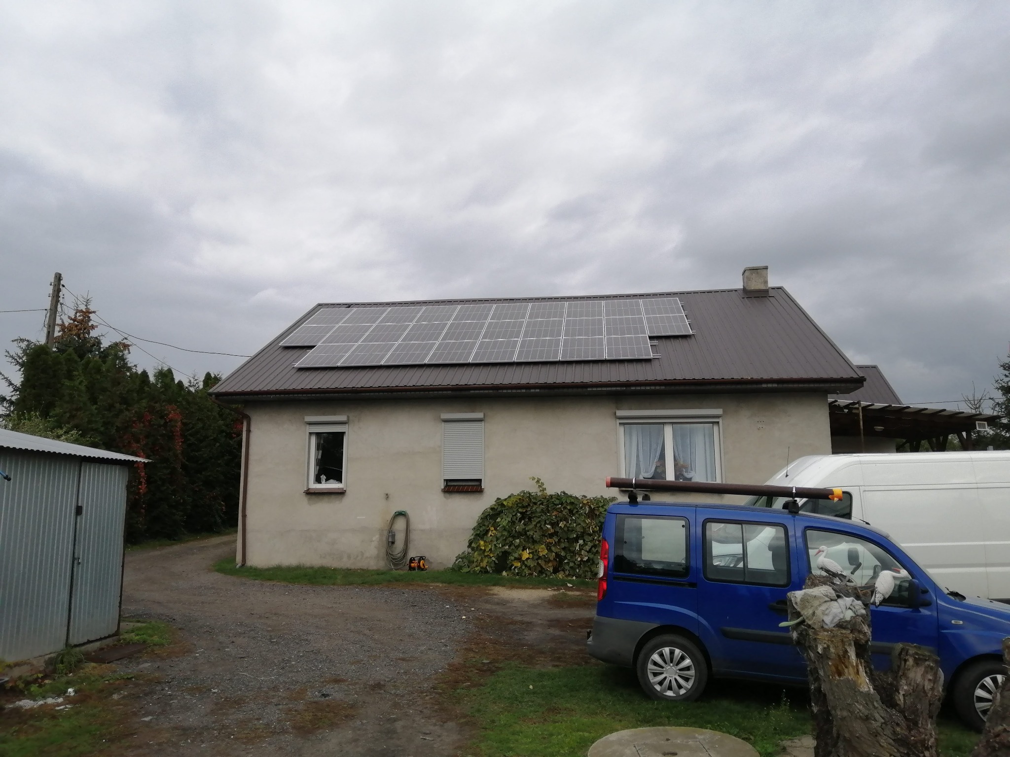 Dom jednorodzinny z brązowym dachem, na którym zamontowano panele słoneczne; na podwórku zaparkowany niebieski samochód z atrapą bocianów na masce.