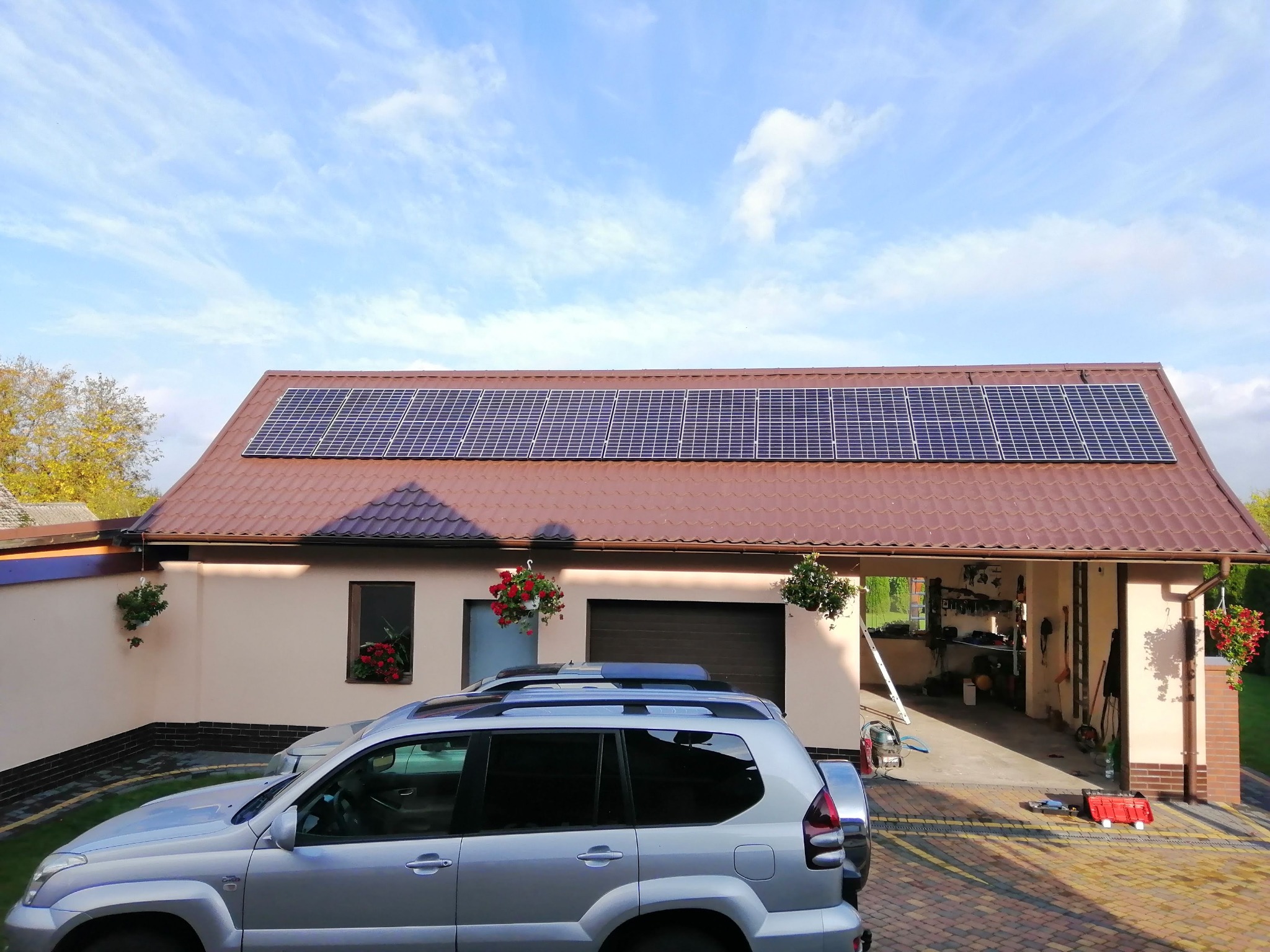 Dom jednorodzinny z brązowym dachem, na którym zamontowane są panele słoneczne, z zaparkowanym srebrnym samochodem terenowym na podjeździe z kostki brukowej.