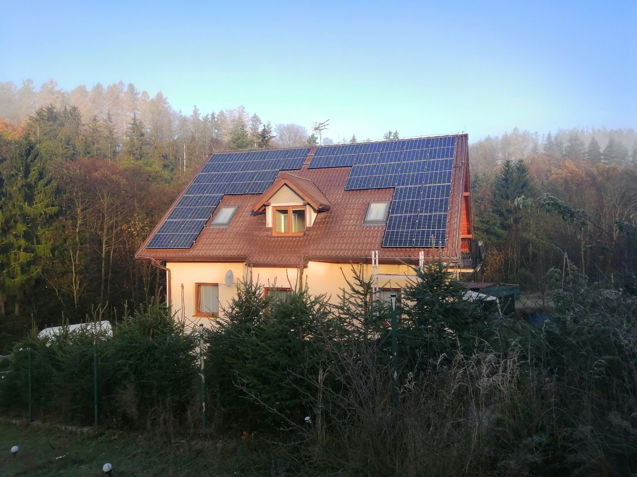 Dom jednorodzinny z panelami fotowoltaicznymi na dachu, otoczony drzewami w jesiennej scenerii z mglistym tłem.