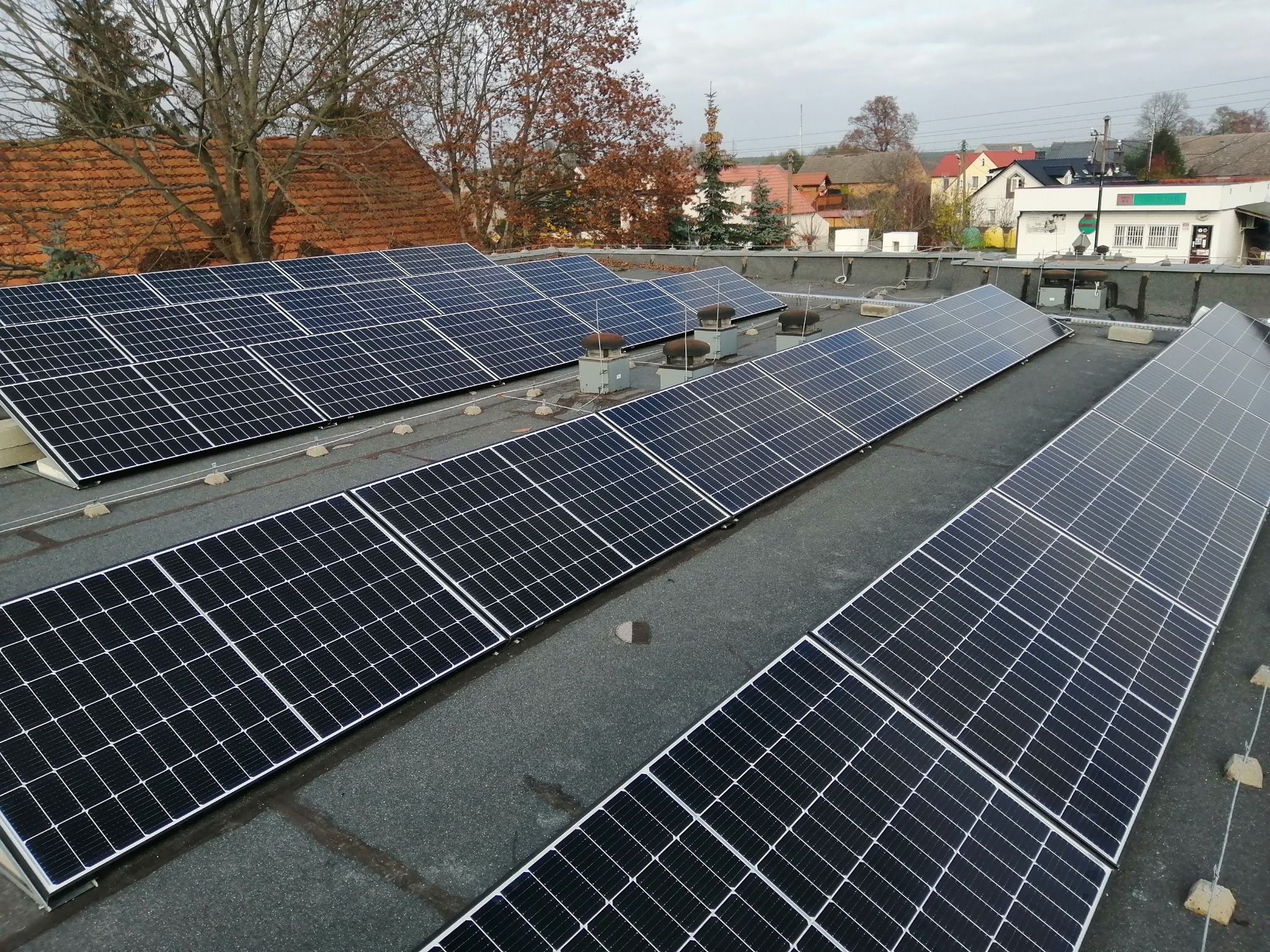 Rząd paneli słonecznych na płaskim dachu budynku z widocznymi elementami mocującymi i wentylacją, w tle zabudowa wiejska pod pochmurnym niebem.