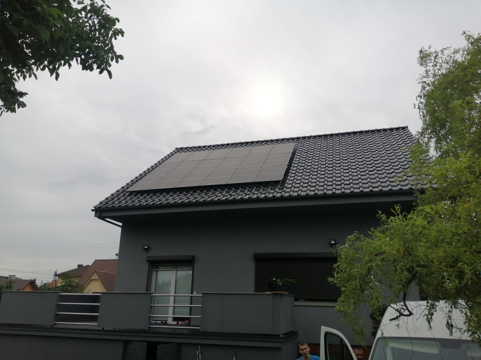 Szary dom jednorodzinny z ciemną dachówką i panelami fotowoltaicznymi na dachu, widoczne fragmenty zieleni drzew i zaparkowany biały samochód dostawczy.