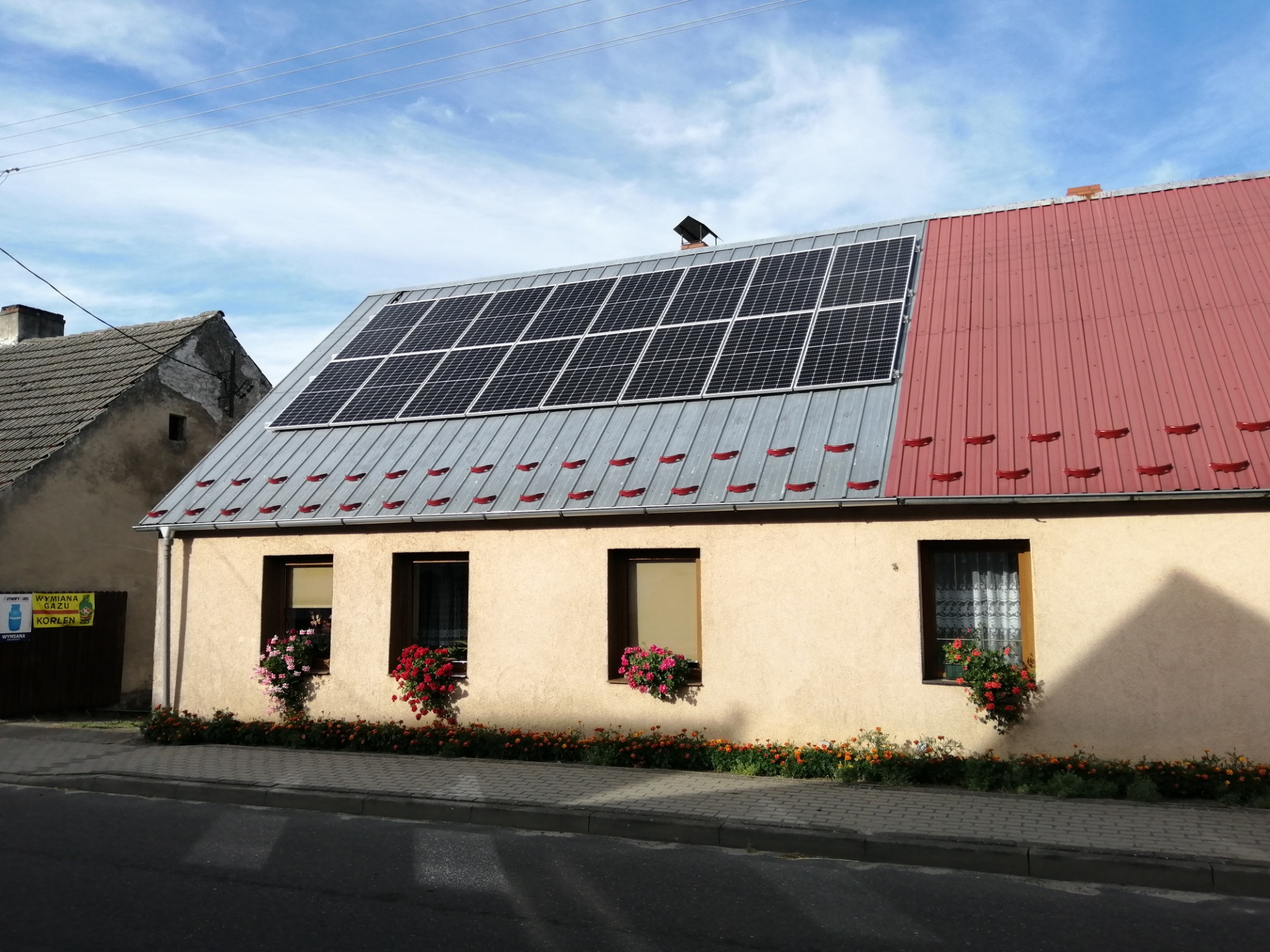Dom jednorodzinny z panelami słonecznymi na dachu, kwiaty w oknach i reklama wymiany gazu obok.