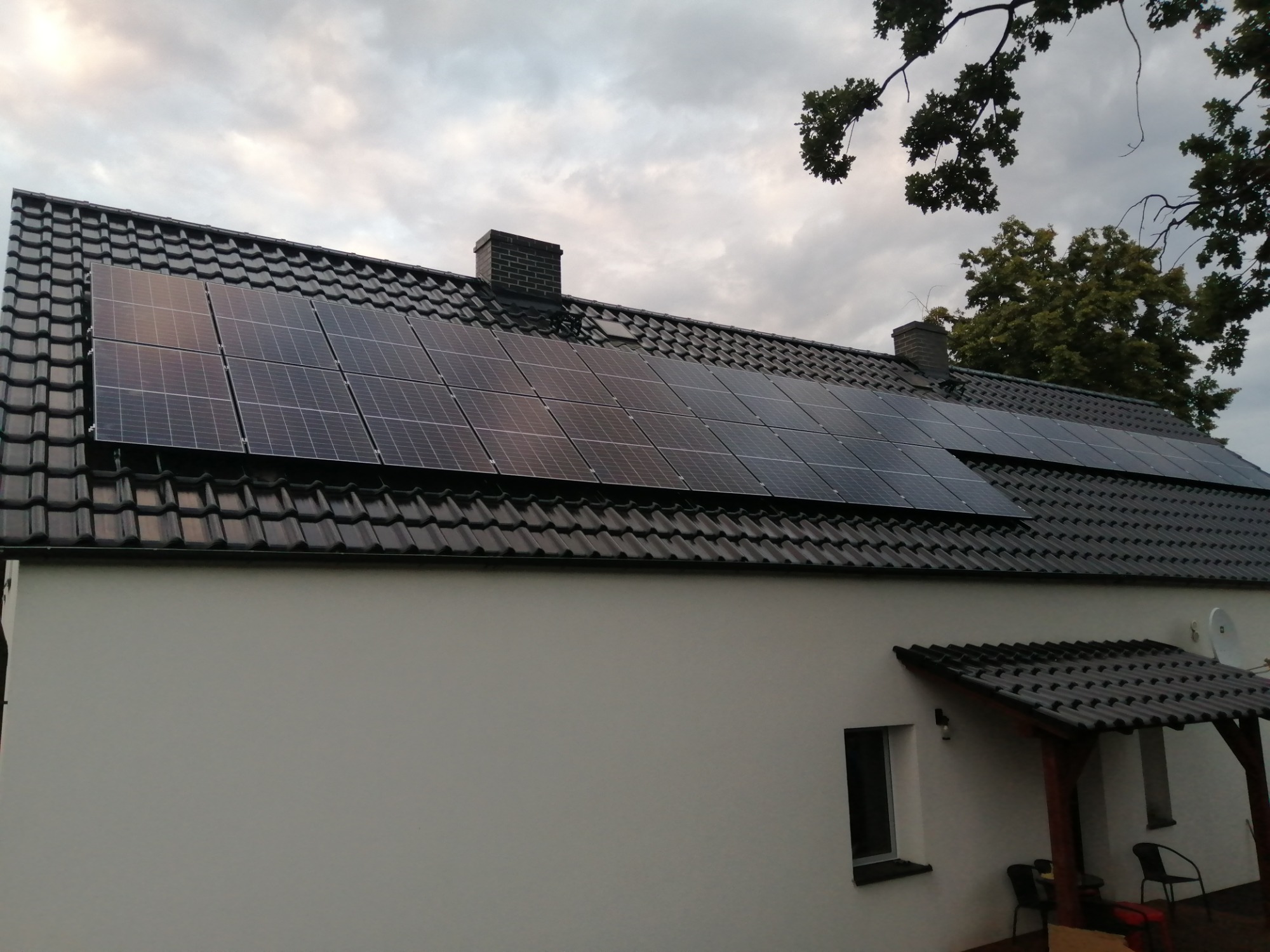 Panele słoneczne zainstalowane na ciemnym, spadzistym dachu domu jednorodzinnego z kominem, widoczne z dołu w pochmurny dzień.