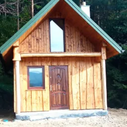 Mały drewniany domek z zielonym dachem, z asymetrycznym oknem na poddaszu i brązowymi drzwiami wejściowymi, otoczony lasem.