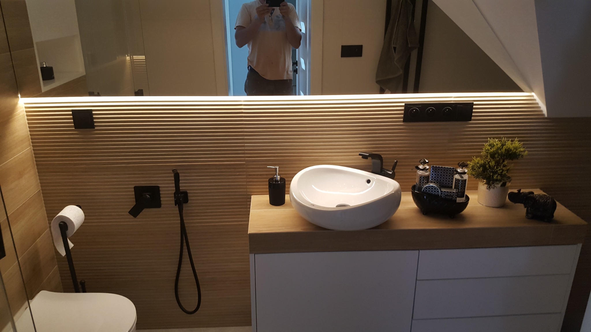 Nowoczesna łazienka w odcieniach beżu z podświetlanym lustrem, umywalką nablatową w kształcie łzy i czarną armaturą, widoczne detale dekoracyjne i toaleta z bidetką.