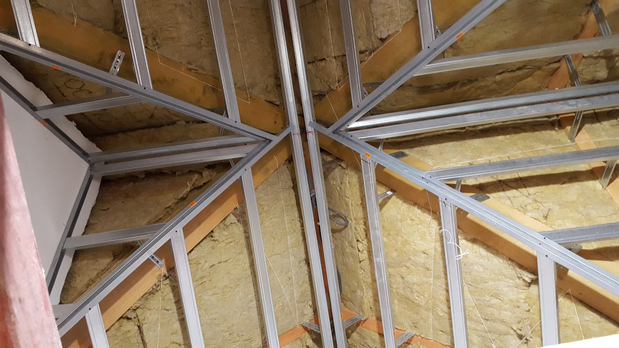 Szkielet sufitu podwieszanego z profili metalowych, wypełniony wełną mineralną, widoczne drewniane elementy konstrukcyjne dachu.