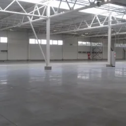 Wnętrze hali przemysłowej z gładką, betonową posadzką, widoczne białe metalowe konstrukcje stropu i słupy wsporcze, okna w górnej części ścian oraz sprzęt i drabina w tle.