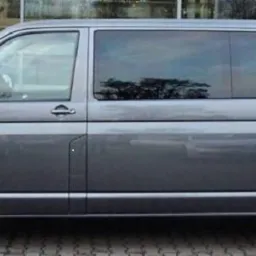 Szary van Volkswagen Transporter T6 z przyciemnianymi szybami, widok z boku na tle budynku z witryną.