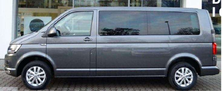 Szary van Volkswagen Transporter T6 z przyciemnianymi szybami, widok z boku na tle budynku z witryną.