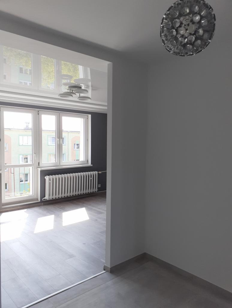 Puste, odświeżone mieszkanie z szarymi ścianami i drewnianą podłogą, widok na pokój z grzejnikiem i oknem, nowoczesna lampa sufitowa.