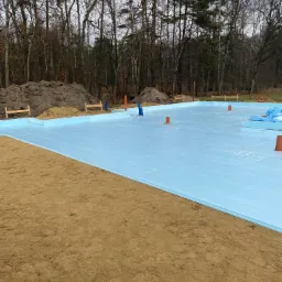 Przygotowany teren pod wylewkę fundamentów z rozłożonym styropianem izolacyjnym, widoczne wystające rury instalacyjne i drewniane znaczniki, w tle drzewa.