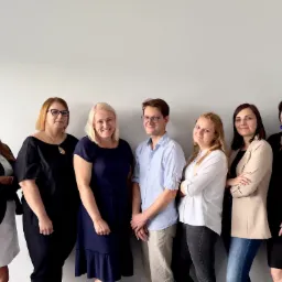 Grupa siedmiu uśmiechniętych osób, głównie kobiet, stojących ramię w ramię na tle jasnoszarej ściany, w różnorodnych strojach biznesowych.