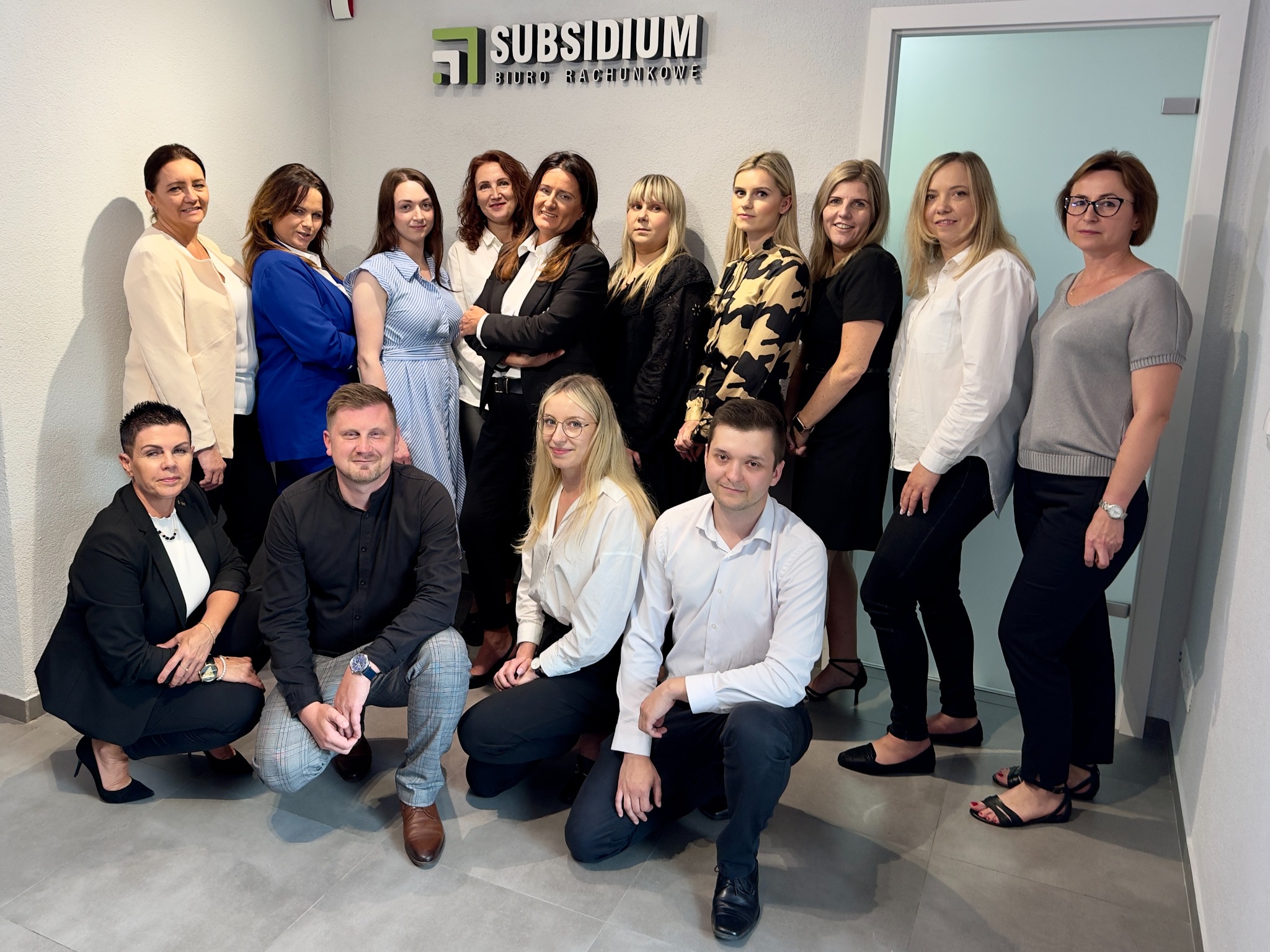 Grupowe zdjęcie pracowników biura rachunkowego Subsidium, część osób stoi, a część klęczy, wszyscy uśmiechnięci, w tle logo firmy na szarej ścianie i białe drzwi.