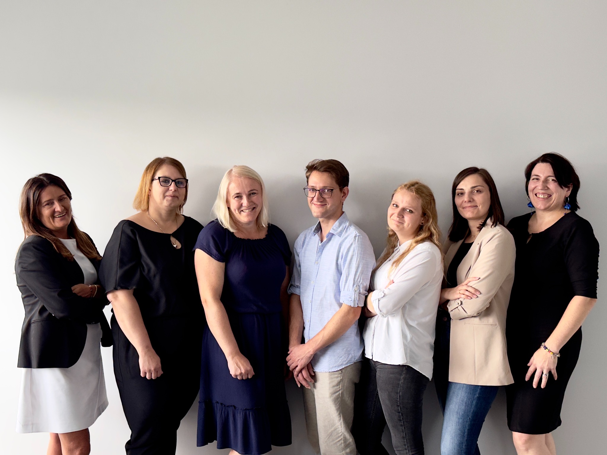 Grupa siedmiu uśmiechniętych osób, głównie kobiet, stojących ramię w ramię na tle jasnoszarej ściany, w różnorodnych strojach biznesowych.