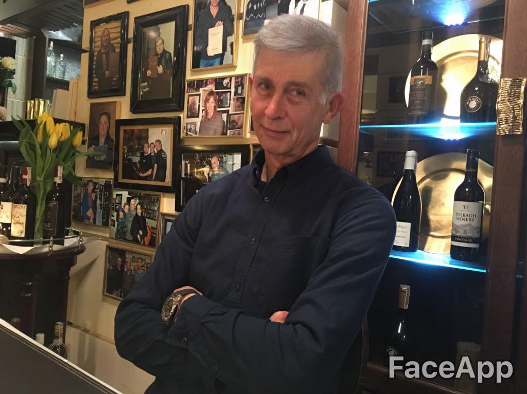 Portret mężczyzny na tle ściany z ramkami i półki z butelkami wina, w restauracji. Uśmiechnięty, z założonymi rękami, w ciemnej koszuli.