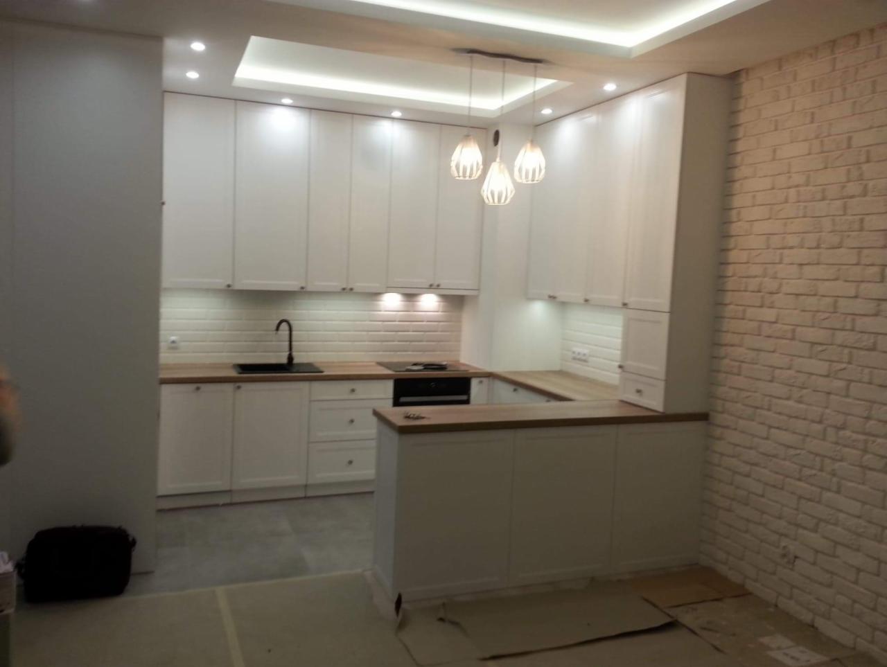 Jasna kuchnia z białymi szafkami, drewnianym blatem i ceglaną ścianą, oświetlona wiszącymi lampami i punktowymi światłami w suficie.