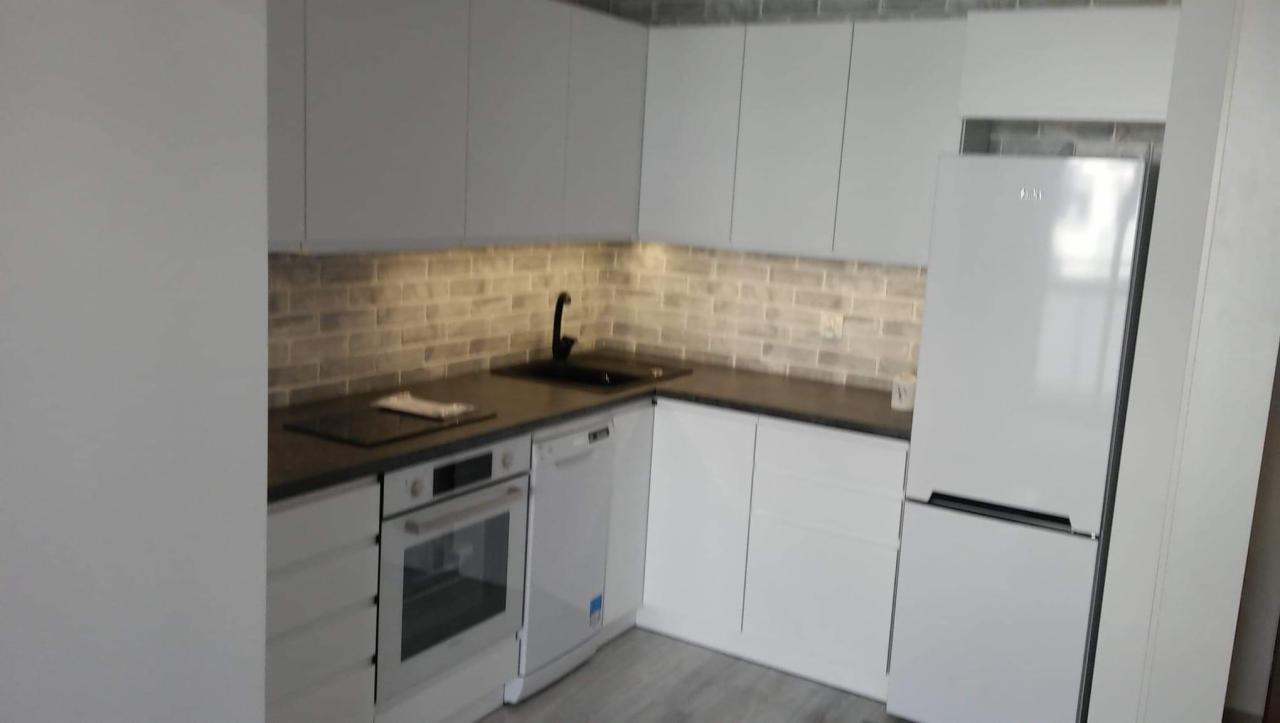 Nowoczesna kuchnia z białymi frontami szafek, ciemnym blatem i ceglaną ścianą nad blatem, oświetlona listwą LED pod szafkami wiszącymi. Widoczny zlew z czarną baterią, piekarnik, zmywarka i lodówka.