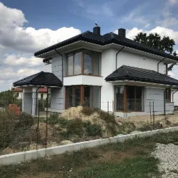 Nowoczesny dom jednorodzinny z białymi ścianami i ciemną dachówką, częściowo wykończony, widoczny zza ogrodzenia na tle pochmurnego nieba. Na kominie siedzi ptak.