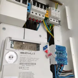 Wnętrze skrzynki elektrycznej z zamontowanym licznikiem energii Smart EMU 3, zabezpieczeniami nadprądowymi Eaton i ogranicznikami przepięć, widoczne przewody elektryczne w kolorach czarnym...