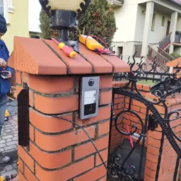 Montaż wideodomofonu na ceglanym słupku ogrodzenia, widoczne narzędzia: śrubokręt i multimetr, w tle dom jednorodzinny.