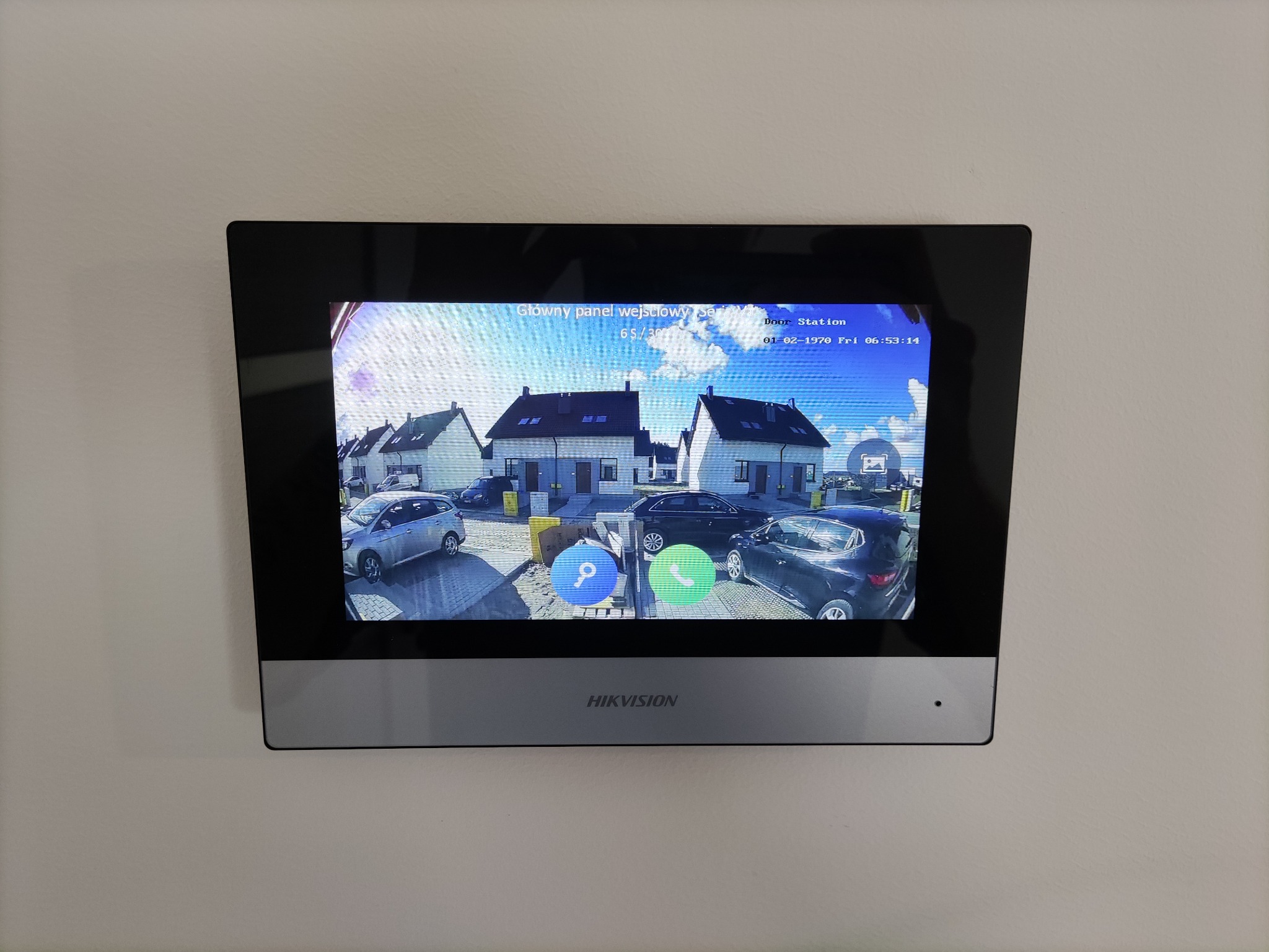 Ekran wideodomofonu Hikvision zamontowanego na białej ścianie, wyświetlający obraz z kamery na zewnątrz, ukazujący domy i zaparkowane samochody w słoneczny dzień.