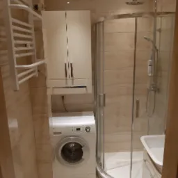 Nowoczesna łazienka z kabiną prysznicową, pralką pod szafką i grzejnikiem drabinkowym, wykończona beżowymi płytkami.