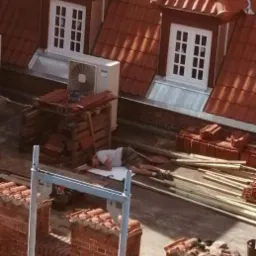 Pracownik w krótkich spodenkach i koszulce leżący na dachu z czerwonej dachówki obok sterty nowych dachówek i klimatyzatora, widoczne okna mansardowe i rusztowanie.