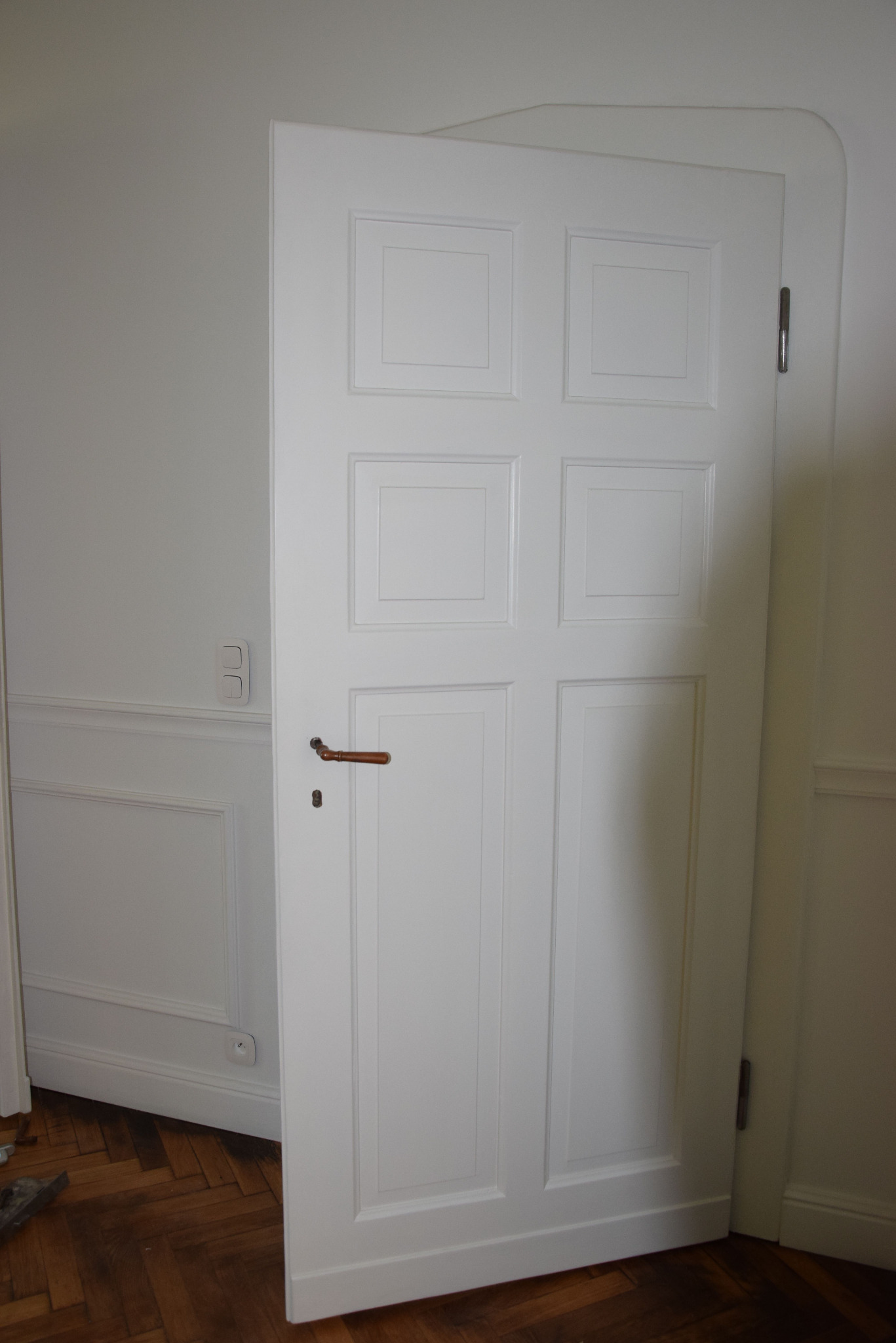 Białe, częściowo otwarte drzwi wewnętrzne z drewnianą klamką i panelami dekoracyjnymi na ścianie, na tle parkietu.