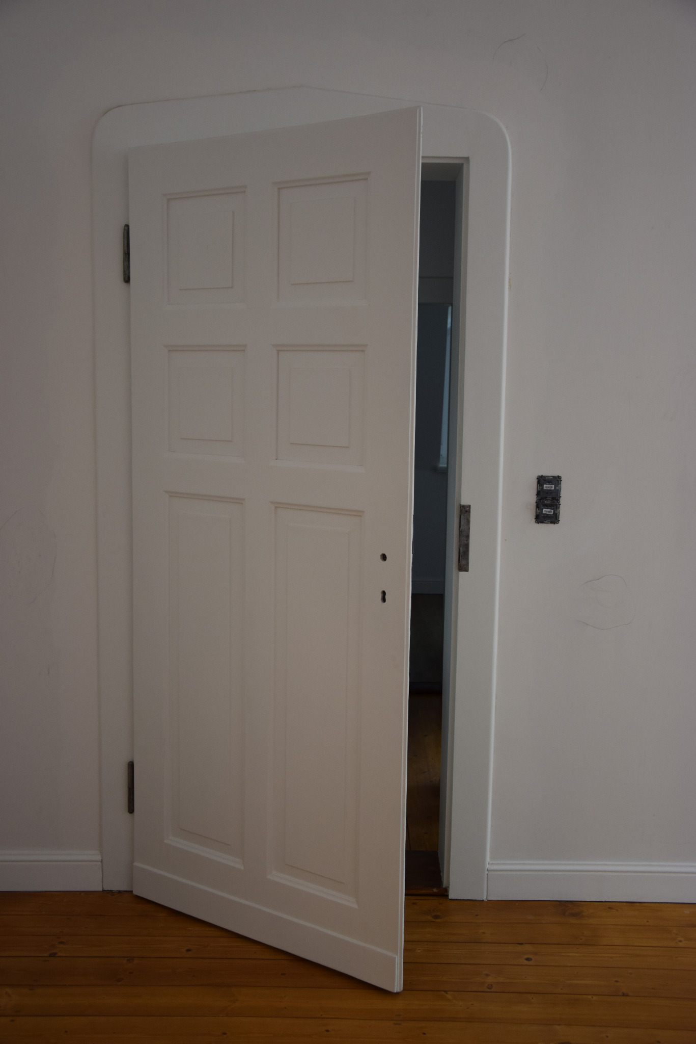 Białe, częściowo otwarte drzwi wewnętrzne z sześcioma panelami, widoczne na tle drewnianej podłogi i białych ścian z widocznymi śladami po naprawach i gniazdkiem elektrycznym.