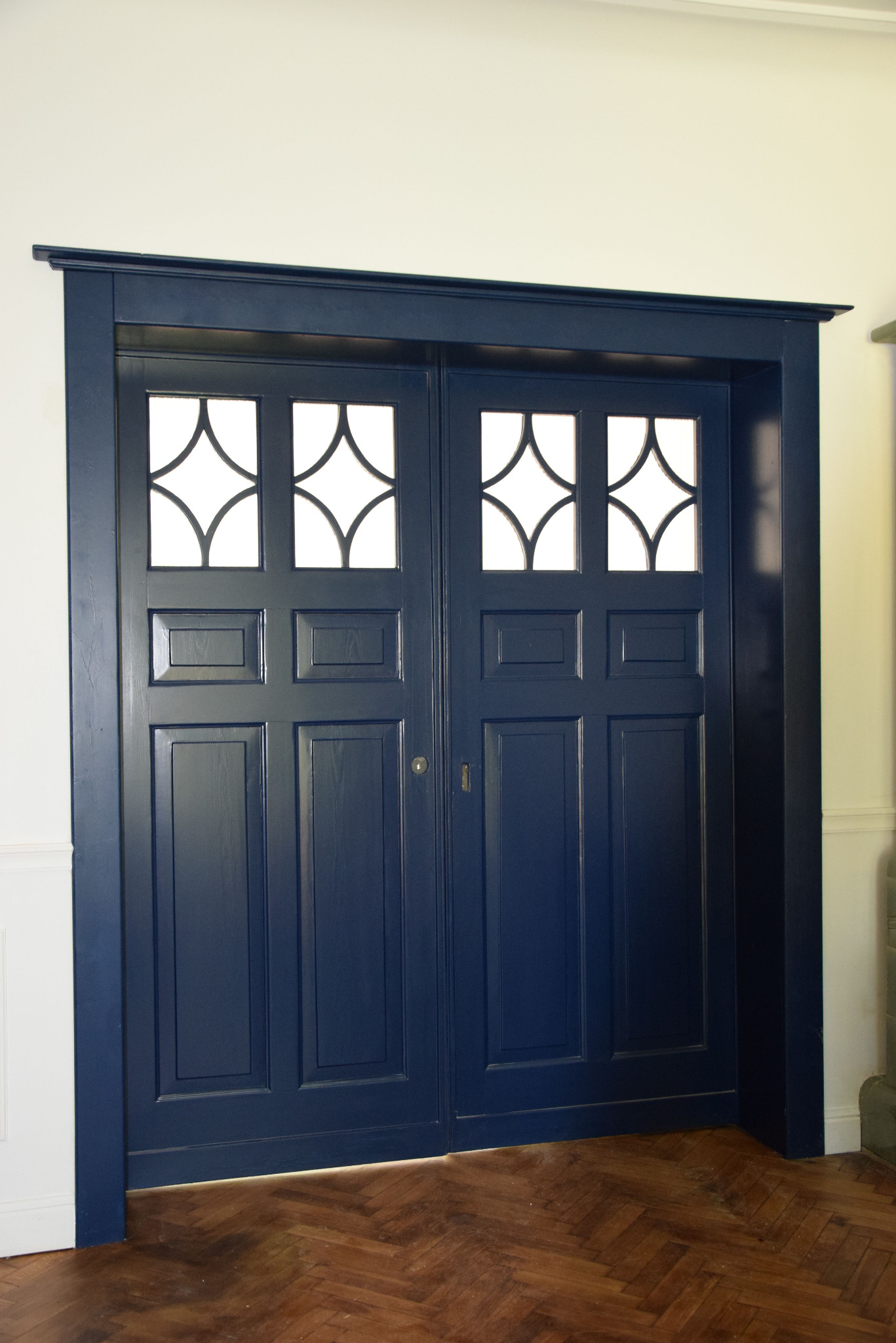 Dwuskrzydłowe, ciemnoniebieskie drzwi wewnętrzne z przeszklonymi, geometrycznymi zdobieniami w górnej części, zamontowane w białej ścianie z widoczną podłogą parkietową.