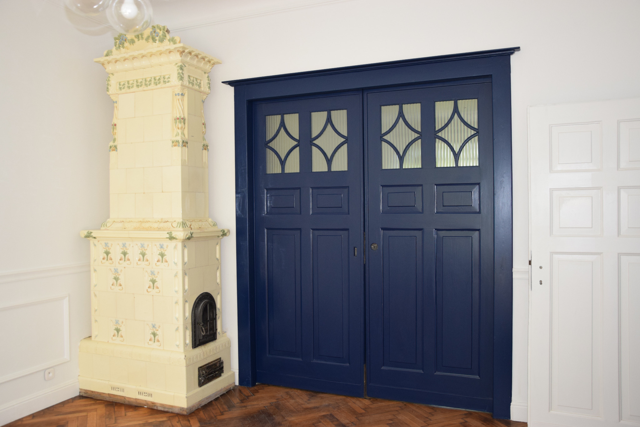 Granatowe, dwuskrzydłowe drzwi przesuwne z szybkami o geometrycznym wzorze, obok biały piec kaflowy z dekoracjami kwiatowymi i białe drzwi w jasnym wnętrzu.