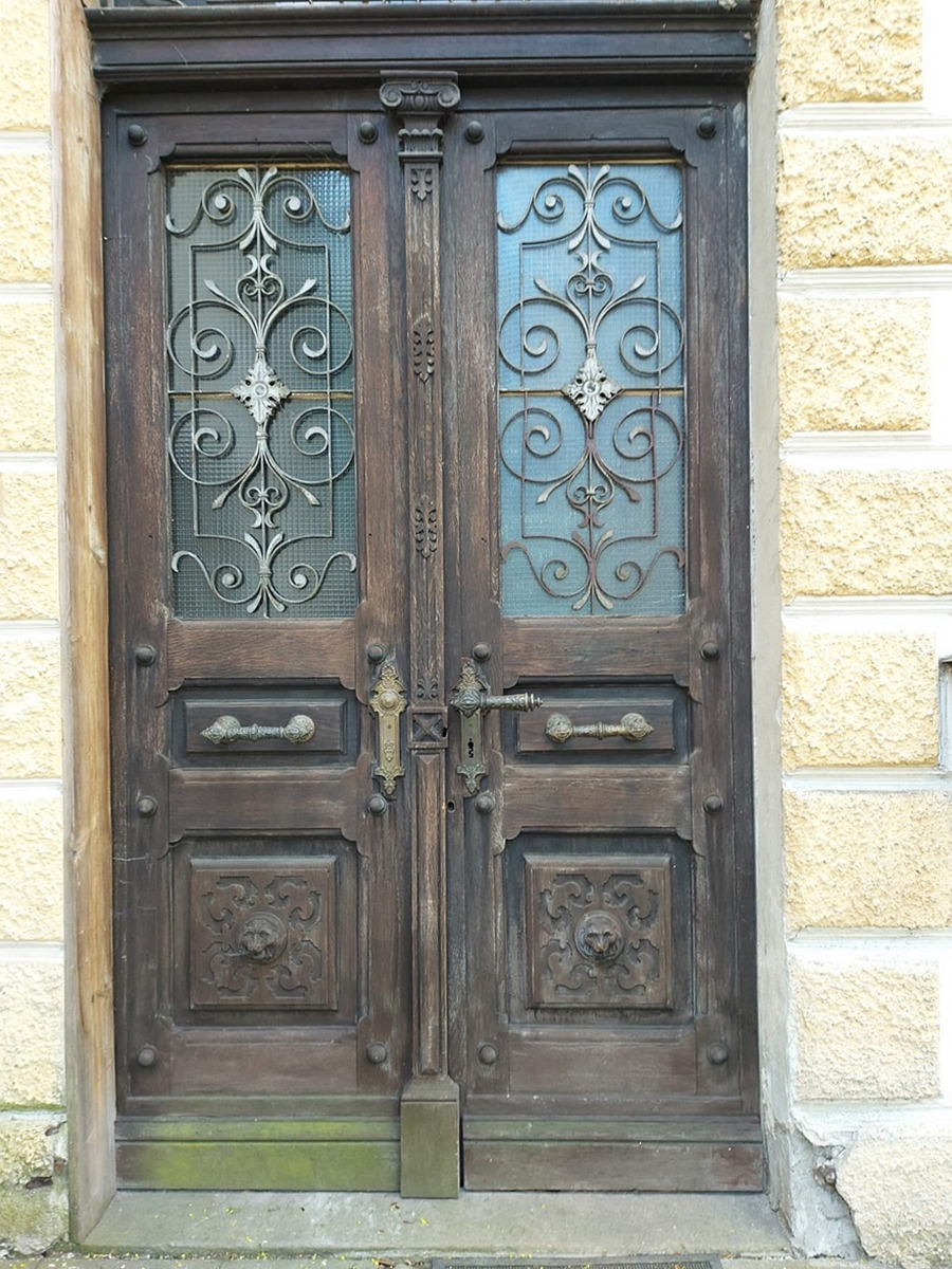 Stare, dwuskrzydłowe drzwi wejściowe z ciemnego drewna, z dekoracyjnymi metalowymi kratami w górnej części i płaskorzeźbami lwich głów w dolnej części, osadzone w murze z jasnego kamienia. Widoczne...