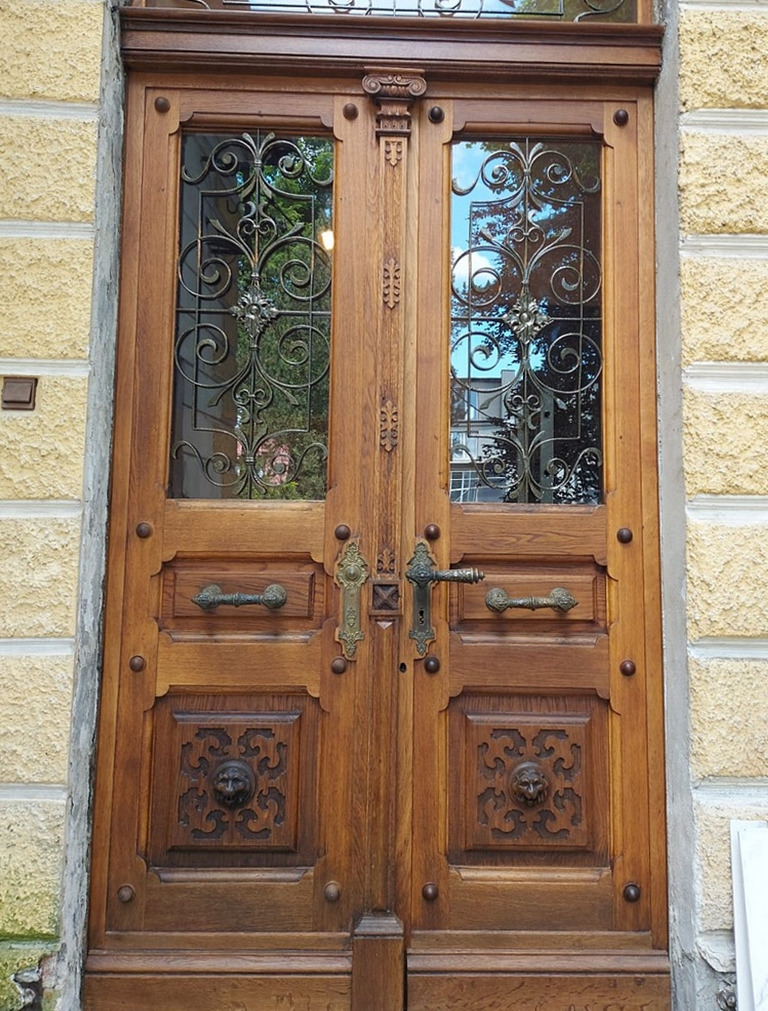 Dwuskrzydłowe, drewniane drzwi wejściowe z dekoracyjnymi metalowymi kratami w górnej części i rzeźbionymi motywami lwów w dolnej części, osadzone w kamiennej fasadzie.