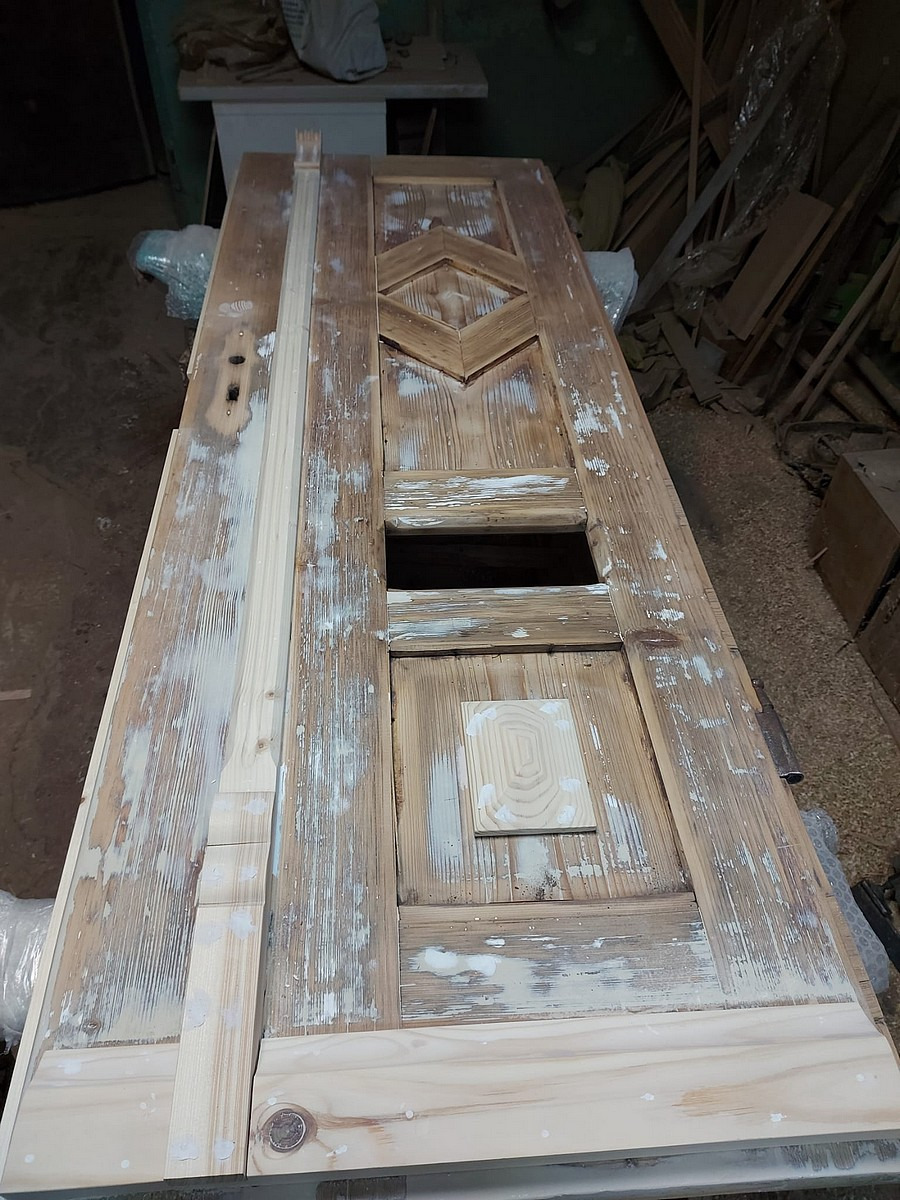 Drewniane drzwi w trakcie renowacji z widocznymi fragmentami starej farby, leżące na warsztatowym stole, z elementami dekoracyjnymi i przygotowanymi listwami do montażu.