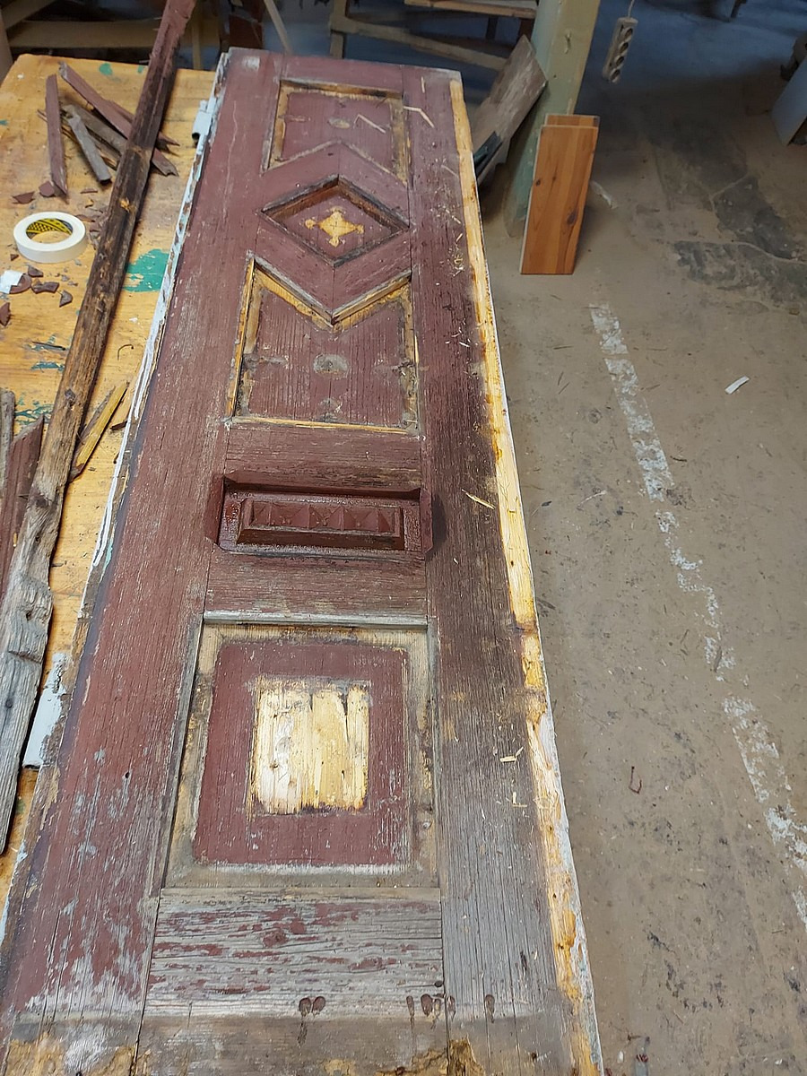 Stara, drewniana, brązowa drzwi z dekoracyjnymi elementami leżąca na warsztatowym stole w trakcie renowacji, z widocznymi śladami farby, uszkodzeniami i fragmentami drewna.