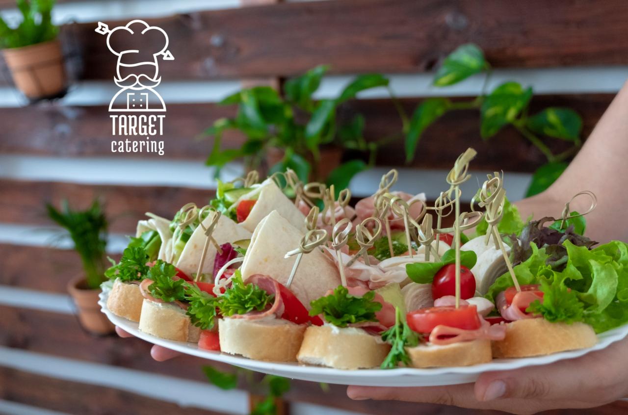 Talerz z różnorodnymi przekąskami cateringowymi, w tym mini kanapeczki i tortille, udekorowany zielonymi roślinami w tle, prezentowany przez osobę.
