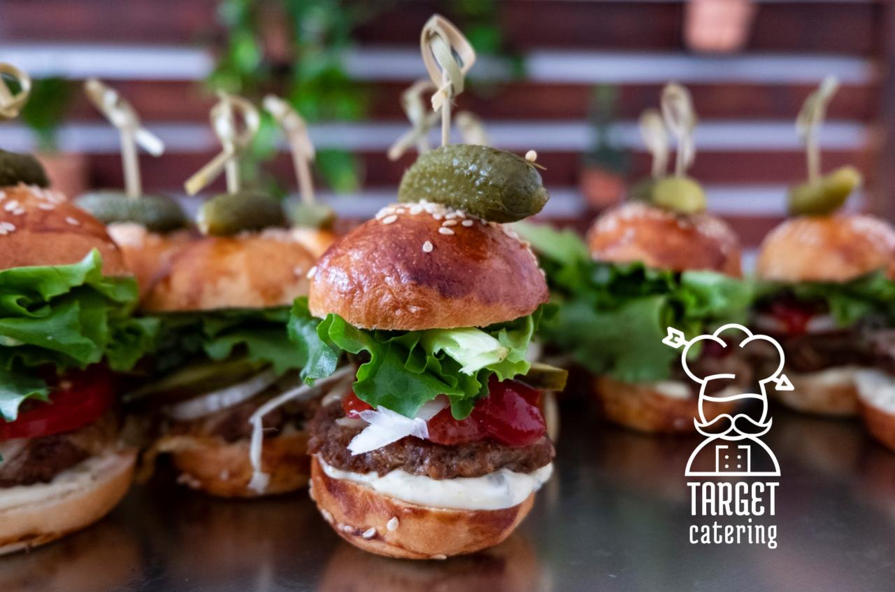 Mini burgery z piklami na wykałaczkach, starannie ułożone na metalowej powierzchni, z logo firmy cateringowej w prawym dolnym rogu.