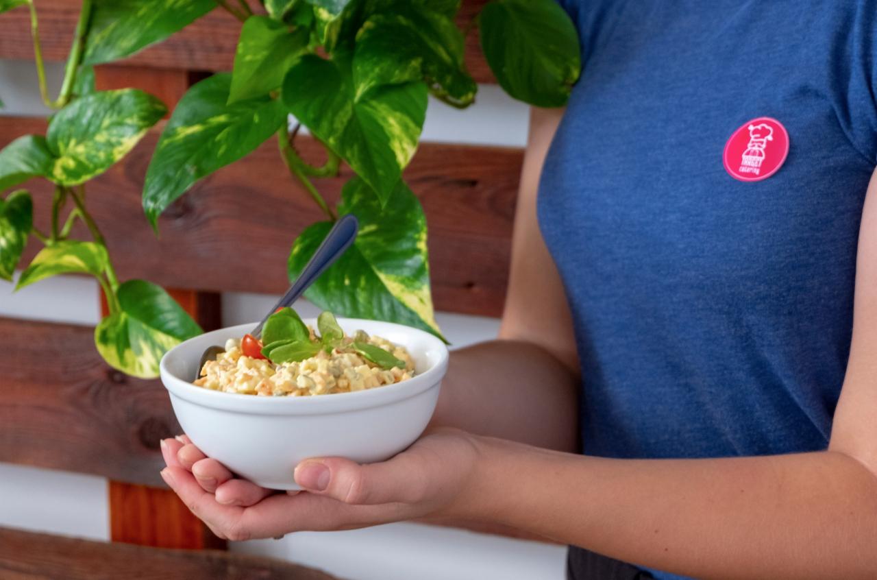 Osoba trzyma białą miskę sałatki jarzynowej z pomidorkiem cherry i bazylią, na tle drewnianej ściany i zielonej rośliny; na koszulce widoczne logo firmy cateringowej.